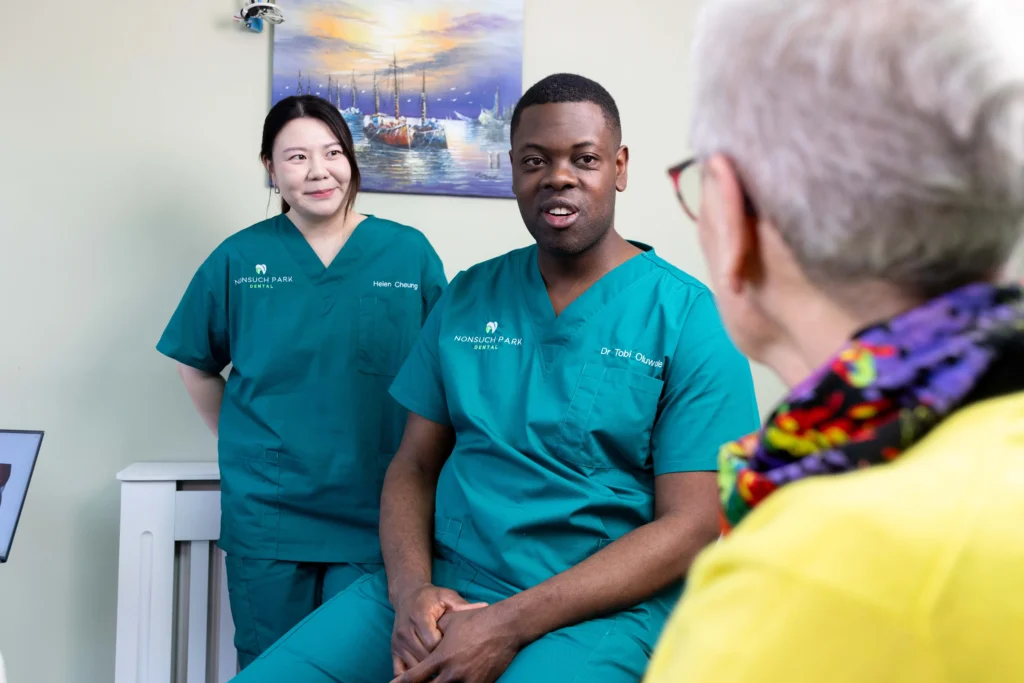 A dental consultation featuring Dr. Tobl Okwuile and Helen Cheung, both in teal scrubs, engaging with a patient.